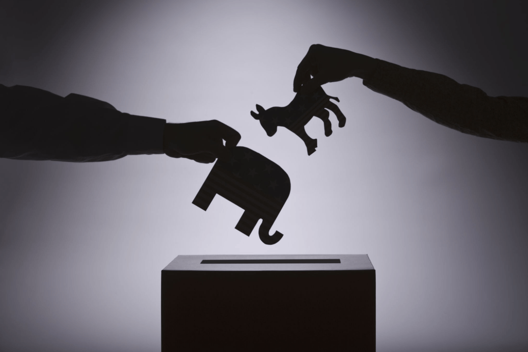 one hand dropping an elephant into a ballot box and one hand dropping a donkey